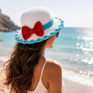 May include: A white crochet sun hat with a red bow and blue trim. The hat is worn by a person looking out at the sea. The hat has a wide brim, ideal for a sunny day at the beach.