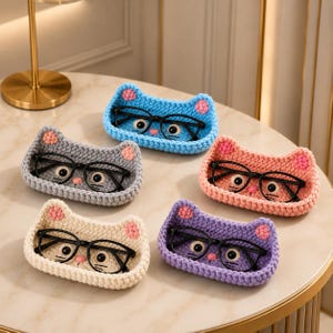 Può includere: Cinque piccoli vassoi all'uncinetto di forma ovale progettati come facce di gatto, ognuno contenente un paio di occhiali con montatura nera. I vassoi sono blu, grigi, color pesca, crema e viola, con orecchie rosa e baffi ricamati.