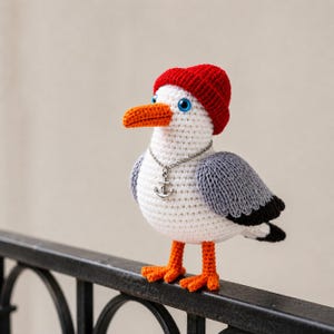 Peut inclure: Un goéland crocheté à la main, portant un bonnet rouge, un bec et des pattes oranges, et des yeux bleus. Le goéland porte un collier argenté avec un pendentif en forme d'ancre. Les ailes sont grises et noires. Il est posé sur une rampe en métal noir.