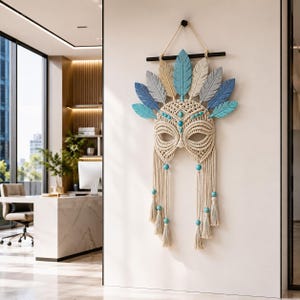 May include: A macrame wall hanging featuring a central mask design made of cream-coloured cord, accented with turquoise beads. The top is adorned with a fan of wooden feathers in shades of blue, teal, and beige, suspended from a black horizontal rod.