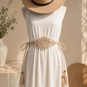 Puede incluir: Un cinturón de macramé beige con un diseño central en forma de diamante y lazos largos con borlas. El cinturón se lleva sobre un vestido blanco, y las borlas tienen cuentas de madera. Un sombrero color canela está encima del maniquí.