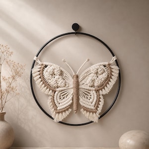 May include: A macrame butterfly wall hanging. The butterfly is crafted with cream and tan-colored rope, and is mounted on a black metal ring. The butterfly's wings have a detailed, textured design. The wall hanging is a decorative piece.