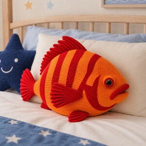 May include: An orange and red crocheted fish cushion with striped detailing and fins. The fish is placed on a bed with a blue star-shaped cushion and a blue and white patterned bedspread. The cushion is a decorative item.