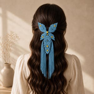 Op de afbeelding: Een lichtblauw macramé haaraccessoire met een bladvormig ontwerp en gele kralen die in de gevlochten koordstructuur zijn verwerkt. Het stuk bevat een lange, bungelende franje van dunne blauwe koorden die naar beneden hangen.