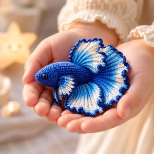 May include: A handcrafted blue and white crochet Betta fish, featuring detailed fins and a dark eye. The fish is held in open hands, highlighting the intricate craftsmanship of the aquatic-themed art piece.