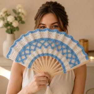May include: A handheld folding fan featuring a light blue and cream crochet lace cover with a lattice pattern. The fan is constructed with light-colored wooden ribs that radiate from the base, which is finished with a small matching cream tassel.