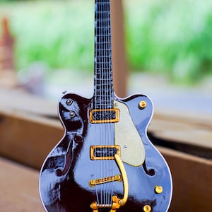 Puede incluir: Una guitarra eléctrica en miniatura con un acabado burdeos oscuro y herrajes dorados. La guitarra tiene un diseño clásico con un golpeador blanco y se exhibe sobre una superficie de madera. La guitarra mide aproximadamente 30 cm de largo.