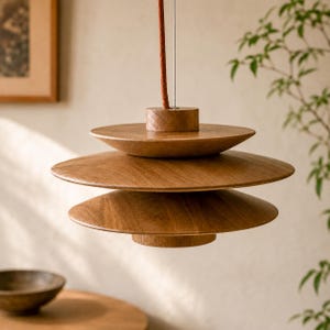 May include: A pendant light fitting featuring three stacked, circular wooden discs with a natural brown grain finish. The fitting is suspended by a single, thin cord with a textured, reddish-brown fabric covering, connecting to a cylindrical wooden top.