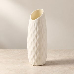 May include: A tall, off-white ceramic vase with a cylindrical, slightly tapered shape. The exterior surface features a repeating, vertical, wave-like textured pattern. The top opening is cut at an angle, revealing a smooth, hollow interior.