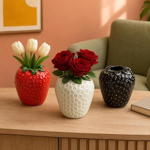 May include: Three ceramic strawberry-shaped vases in red, white, and black. The red vase holds three white tulips, the white vase holds three red roses, and the black vase is empty. Each vase features a textured surface with green leaves at the top.