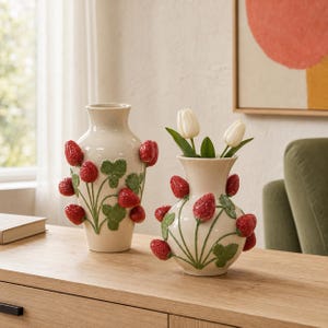 May include: Two cream-coloured ceramic vases featuring raised, three-dimensional red strawberry motifs and green leaves. The taller vase has a slender neck, while the shorter, rounder vase contains two white tulip flowers emerging from the top.