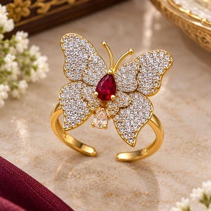 May include: A gold-toned ring featuring a butterfly design encrusted with numerous small clear stones. The centre of the butterfly displays a prominent pear-shaped red stone, while a smaller clear pear-shaped stone sits just below it.