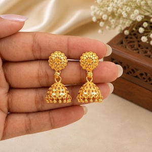 Può includere: Un paio di orecchini jhumka dorati con un perno circolare caratterizzato da intricati motivi floreali in filigrana e un pendente a forma di campana decorato con fasce orizzontali testurizzate e piccole perline pendenti sul bordo inferiore.