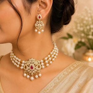 Puede incluir: Un collar tipo gargantilla en tono dorado y pendientes a juego. El collar presenta tres hileras de perlas blancas con un colgante central dorado incrustado con piedras claras y una piedra roja en forma de lágrima, con perlas colgantes.
