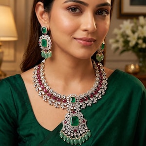 May include: A jewelry set consisting of a necklace and matching earrings. The necklace features a large central emerald-cut green stone, surrounded by white crystals, red stones, and dangling light green beads. Earrings mirror this design.