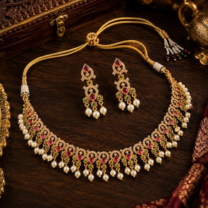May include: A gold-toned necklace and matching earring set featuring intricate metalwork, embedded with small clear stones, vibrant red and green accents, and dangling white pearls. The necklace includes an adjustable cord with pearl-tipped ends.