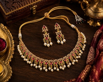 Puede incluir: Un conjunto de collar y pendientes en tono dorado con intrincados detalles metálicos, incrustaciones de pequeñas piedras claras, acentos en rojo y verde vibrante y perlas blancas colgantes. El collar incluye un cordón ajustable con perlas en los extremos.