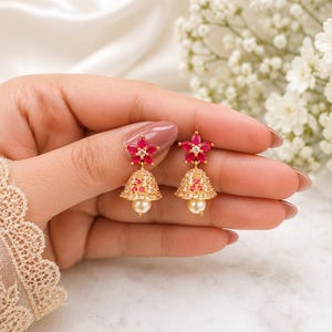 Puede incluir: Un par de pendientes jhumka dorados con un poste en forma de flor de cinco pétalos con piedras rosas vibrantes y un cristal central. El colgante en forma de campana está incrustado con piedras claras, detalles rosas y una perla blanca colgante.