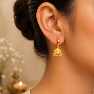 Könnte beinhalten: Ein goldener Jhumka-Ohrring mit einem polierten Creolen-Verschluss, der mit einem glockenförmigen Anhänger verbunden ist, welcher filigrane Metallarbeiten und kleine hängende Perlen am unteren Rand aufweist, alles in einem hellen Gelbgoldton.