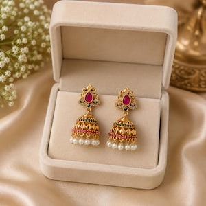 Könnte beinhalten: Ein Paar goldfarbene Jhumka-Ohrringe mit einem tropfenförmigen rosa Stein an der Oberseite, filigraner Metallarbeit, kleinen grünen Akzenten und einer Reihe hängender weißer Perlen an der Basis, präsentiert in einer beigen Samtschmuckschatulle.