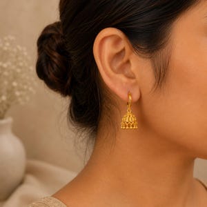 Könnte beinhalten: Ein goldener Jhumka-Ohrring bestehend aus einem kleinen Ring, der an einem glockenförmigen Anhänger mit filigraner Metallarbeit und einer Reihe winziger, hängender Perlen am unteren Rand befestigt ist und am Ohrläppchen getragen wird.