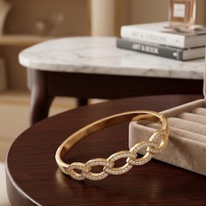 Puede incluir: Un brazalete rígido articulado de tono dorado que presenta una serie de eslabones ovalados entrelazados. La sección frontal del brazalete está adornada con pequeñas piedras transparentes engastadas en pavé que captan la luz, creando un efecto brillante.