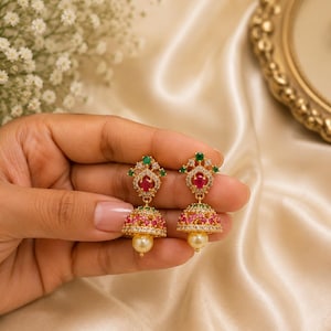Peut inclure: Une paire de boucles d'oreilles jhumka dorées avec un design en forme de cloche orné de petites pierres roses et blanches. La partie supérieure comprend une pierre rouge ovale centrale, des accents verts et des cristaux, avec une perle crème.