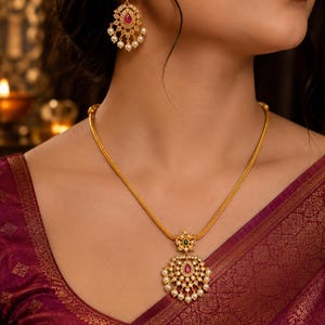 Peut inclure: Un ensemble collier et boucles d'oreilles pendantes doré avec un travail du métal complexe, de petites perles blanches et des pierres centrales en forme de goutte rouge et verte. Le collier possède une fine chaîne texturée et un pendentif rond.