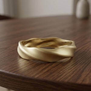 Puede incluir: Un brazalete rígido de tono dorado con un acabado mate suave y un diseño ondulado continuo que crea una serie de curvas suaves y redondeadas alrededor de toda la circunferencia de la banda metálica circular.