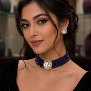May include: A multi-strand dark blue beaded choker necklace featuring a central oval-shaped pendant with a clear stone set in a gold-toned frame, accompanied by matching stud earrings with a similar clear stone and gold-toned border design.