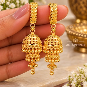 Könnte beinhalten: Ein Paar goldene Jhumka-Ohrringe mit einem blattförmigen Stecker, einer glockenförmigen Kuppel mit filigranem Blumenmuster und mehreren Reihen hängender Goldperlen. Der Text anikjewels ist auf der rechten Seite des Bildes zu sehen.