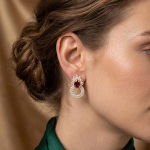 May include: A gold-toned earring featuring a central pear-shaped deep red gemstone surrounded by a halo of small clear stones, with a circular drop design below also encrusted with clear stones, creating a layered and ornate appearance.