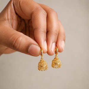 Puede incluir: Un par de pendientes de aro dorados con colgantes en forma de campana jhumka. Cada colgante presenta un intrincado patrón de filigrana calada y está rematado con diminutas cuentas doradas colgantes a lo largo del borde inferior.