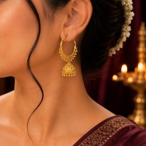 May include: A gold hoop earring featuring a dangling bell-shaped jhumka pendant. The hoop is adorned with small gold beads along the bottom edge, and the bell-shaped charm has a textured, perforated surface with tiny hanging gold spheres.