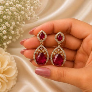 May include: A pair of gold-toned drop earrings featuring a teardrop-shaped deep red gemstone at the base, topped with a smaller matching teardrop stone. Both stones are surrounded by a halo of small, clear, sparkling white accent stones.