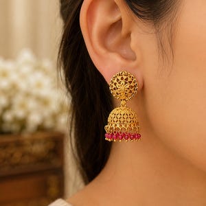 Puede incluir: Un pendiente jhumka de tono dorado con un poste circular de intrincados patrones de filigrana. El colgante en forma de campana presenta un trabajo de metal detallado y termina con una hilera de pequeñas cuentas rosa intenso.