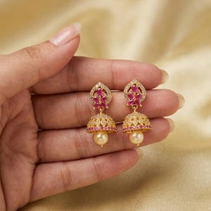 Può includere: Un paio di orecchini jhumka dorati con una parte superiore a goccia decorata con pietre rosa taglio marquise e cristalli trasparenti. La base a campana è impreziosita da cristalli, una fila di pietre rosa e una perla color crema pendente.
