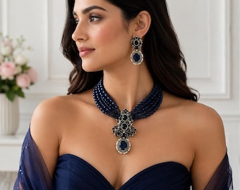 Può includere: Una collana a più fili di perline blu scuro con un ciondolo centrale decorato con pietre blu scuro e piccoli accenti bianchi, abbinata a orecchini pendenti coordinati con una pietra blu scuro circondata da piccole perline bianche.