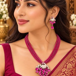 Puede incluir: Un collar de varias hebras con pequeñas cuentas magenta y un colgante central en forma de pavo real con piedras blancas y gemas magenta en forma de lágrima. Los pendientes a juego presentan un motivo de pavo real con piedras blancas.