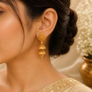 May include: A gold jhumka earring featuring a circular filigree stud at the top, connected to two stacked, bell-shaped tiers with intricate metalwork and a small dangling bead at the bottom, showcasing a polished metallic finish throughout.