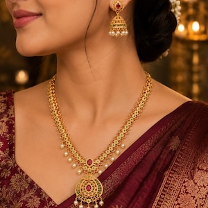 May include: A gold-toned necklace and matching jhumka earring set featuring intricate metalwork, embedded with small red and green stones, and accented with dangling white pearls. The necklace includes a large circular pendant with a central red stone.