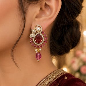 May include: A gold-toned earring featuring a central pear-shaped red stone surrounded by a halo of smaller pink stones and clear crystals. The design includes a white pearl at the top, a cluster of tiny pearls, and a dangling pink oval bead at the base.