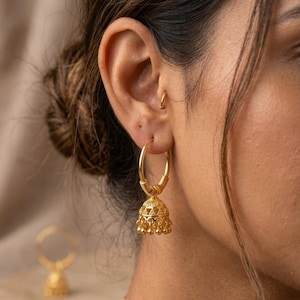 Könnte beinhalten: Ein goldener Creolen-Ohrring mit einem baumelnden, glockenförmigen Jhumka-Anhänger, der mit filigranen Mustern und kleinen kugelförmigen Perlen am unteren Rand verziert ist. Der Ohrring besteht aus einem glatten Ring und einem texturierten Spiraldetail.