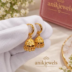 Könnte beinhalten: Ein Paar goldene Jhumka-Ohrringe mit einem polierten Reifen oben, der mit einem glockenförmigen Anhänger verbunden ist, welcher komplizierte Filigranmuster und kleine hängende Goldperlen an der Basis aufweist, präsentiert auf weißem Stoff.