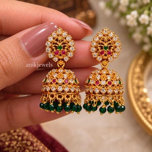 May include: A pair of gold-tone jhumka earrings featuring a teardrop-shaped top and a bell-shaped base, both encrusted with clear white stones, small red accents, and green center stones. The base is finished with dangling green beads. Text reads anikjewels.
