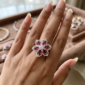 以下が含まれることがあります： 大きな花のデザインが特徴のシルバー調の指輪。中央には小さな透明な石のクラスターがあしらわれ、その周囲を6つのマーキスカットの濃いピンク色の宝石が囲んでいます。花柄のモチーフ全体が小さな透明な石で縁取られています。