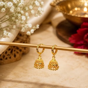 May include: A pair of gold hoop earrings featuring dangling bell-shaped jhumka charms. Each charm has a textured, twisted metal surface and a row of small, spherical beads hanging from the bottom edge, suspended from a polished gold hoop.