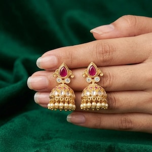 May include: A pair of gold-toned jhumka earrings featuring a teardrop-shaped red stone at the top, a small clear round stone below it, and a bell-shaped base adorned with red stones, clear stones, and a row of small white dangling pearls.