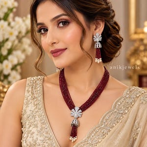 May include: A multi-strand necklace and matching jhumka earrings featuring deep red faceted beads, silver-toned fan-shaped accents encrusted with clear stones, and small white pearl drops. The jewelry set is branded with the text anikjewels.