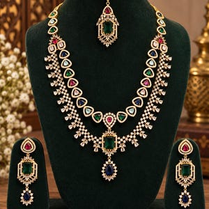 May include: A multi-layered gold-tone necklace set featuring emerald green, sapphire blue, and ruby red teardrop and rectangular stones accented with clear crystals. The set includes matching drop earrings and a matching maang tikka headpiece.
